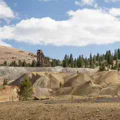 An old wooden mine headframe stands atop a pile of gravel behind several mounds of dirt.