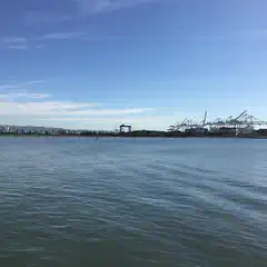 A waterway extends toward a waterfront industrial area with large container cranes and buildings in the distance.