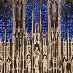 A Gothic facade features intricate stone carvings, tall statues, and vibrant blue stained glass windows arranged in vertical panels.