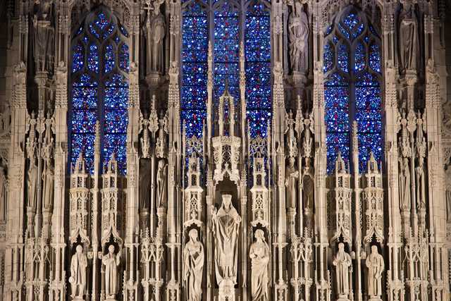 A Gothic facade features intricate stone carvings, tall statues, and vibrant blue stained glass windows arranged in vertical panels.