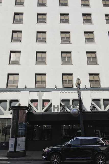 A multistory building with numerous rectangular windows displays a prominent “HOTEL” sign above a dark entrance and a parked black car.