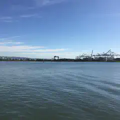 A waterway extends toward a waterfront industrial area with large container cranes and buildings in the distance.