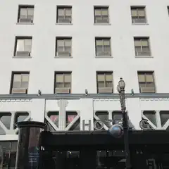 A multistory building with numerous rectangular windows displays a prominent “HOTEL” sign above a dark entrance and a parked black car.