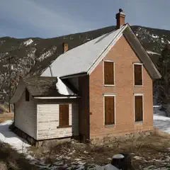 An old two-story house made of red brick has boarded-up windows and is surrounded by snow-covered ground, with mountains visible in the background.