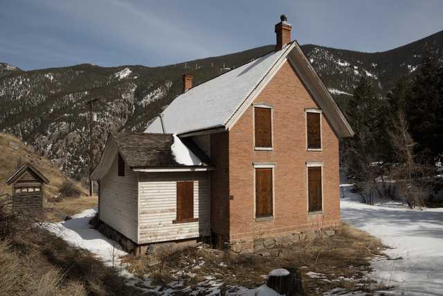 An old two-story house made of red brick has boarded-up windows and is surrounded by snow-covered ground, with mountains visible in the background.