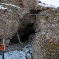A dark cave opening is visible in a rocky hillside, with a warning sign and a wire fence in the foreground.
