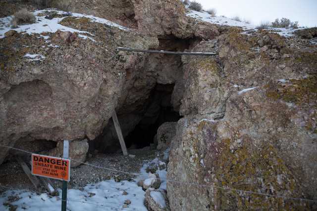 A dark cave opening is visible in a rocky hillside, with a warning sign and a wire fence in the foreground.