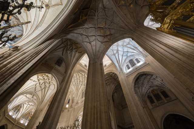 A cathedral interior with tall arched columns, pointed vaults, and stained glass windows allowing natural light to filter through.