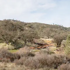 A fenced picnic area with tables and benches sits in a dry, grassy clearing surrounded by sparse trees and rolling hills.