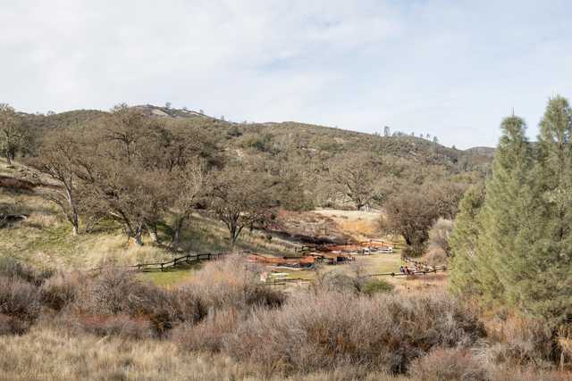 A fenced picnic area with tables and benches sits in a dry, grassy clearing surrounded by sparse trees and rolling hills.