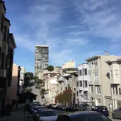 A street lined with parked cars on a hillside cityscape scene featuring many buildings under blue skies with some clouds.