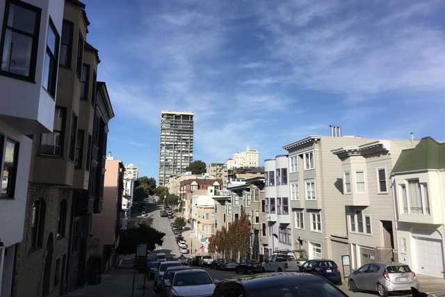 A street lined with parked cars on a hillside cityscape scene featuring many buildings under blue skies with some clouds.