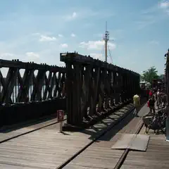 A wooden pier extends toward the water, and a ship's mast is visible in the distance.