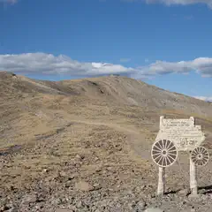 An old wooden sign, shaped like a stage coach, stands on a rocky mountain top with mountains and clouds in the background.