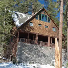 A two-story wooden house with a stone foundation stands among trees and snow.