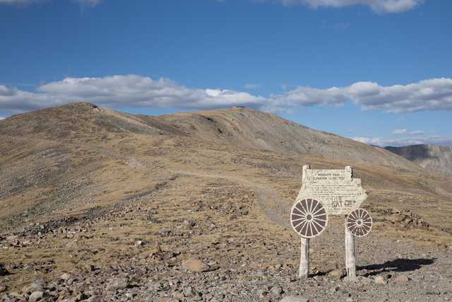 An old wooden sign, shaped like a stage coach, stands on a rocky mountain top with mountains and clouds in the background.