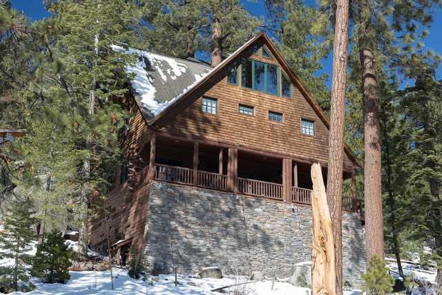 A two-story wooden house with a stone foundation stands among trees and snow.