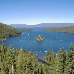 A serene lake with a small island and boats on the water, surrounded by lush green trees under a clear blue sky.