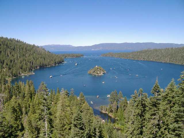 A serene lake with a small island and boats on the water, surrounded by lush green trees under a clear blue sky.