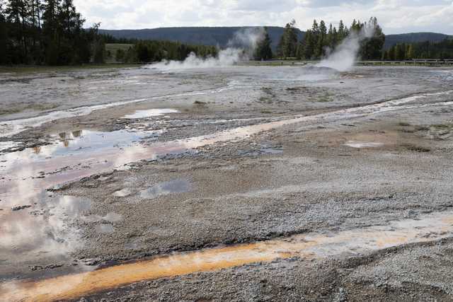 A geothermal area contains cracked earth, steam vents, and pools of water tinted with yellow and brown.