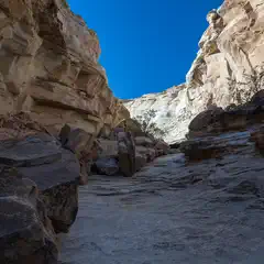 A narrow canyon with tall, layered rock walls and a rocky floor under a clear blue sky.