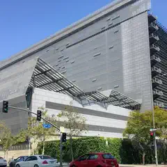An angular, modern building constructed from light-colored stone blocks with metal grating on its roof is visible behind trees and a hedge.