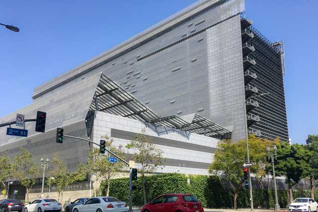 An angular, modern building constructed from light-colored stone blocks with metal grating on its roof is visible behind trees and a hedge.