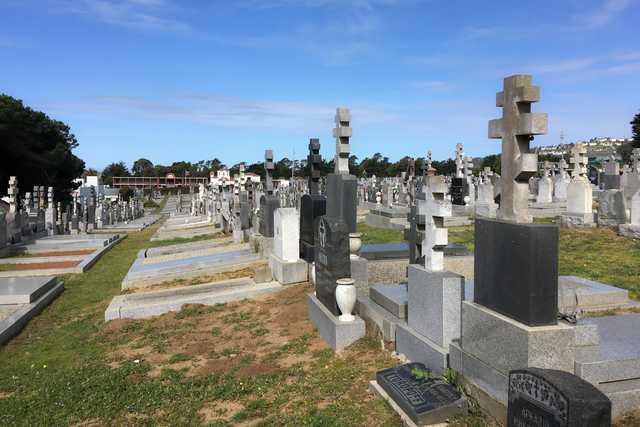 Cemetery with many headstones and grave markers, some decorated with crosses and other symbols, surrounded by trees under a blue sky.