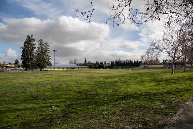 A large, open grassy field under a partly cloudy sky with trees and buildings in the distance.