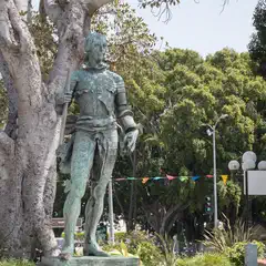 A bronze statue of a man dressed in medieval armor stands on a pedestal near trees and lampposts.
