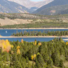 A body of water is surrounded by evergreen trees and distant mountains, with two sailboats visible on the water.