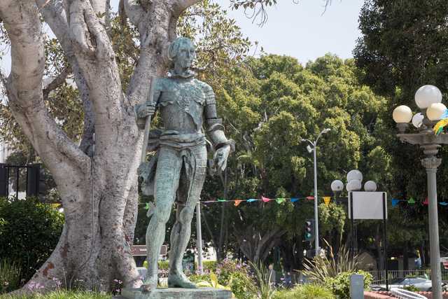 A bronze statue of a man dressed in medieval armor stands on a pedestal near trees and lampposts.