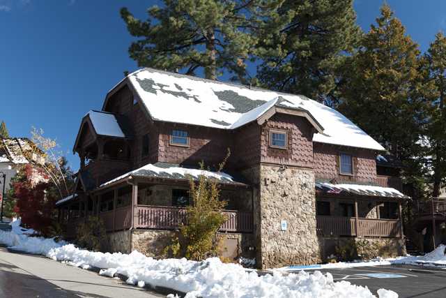 A large wooden lodge with a stone foundation and snow on its roof, surrounded by trees and a parking lot.