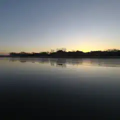 A body of water reflecting sunset colors, with silhouetted trees and buildings on the opposite shore.