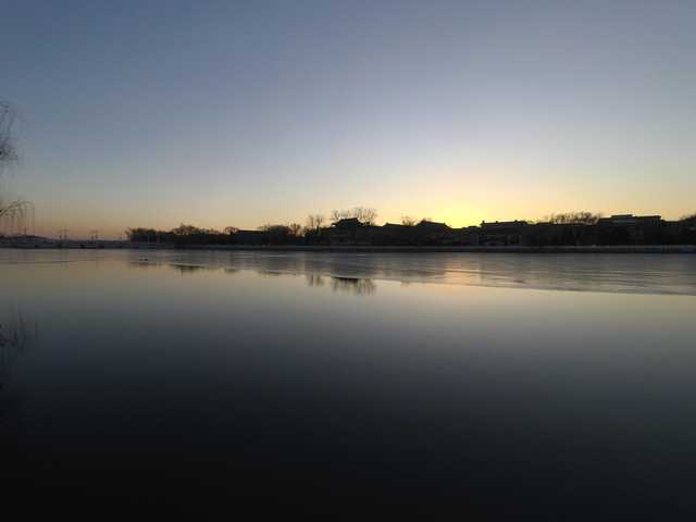 A body of water reflecting sunset colors, with silhouetted trees and buildings on the opposite shore.