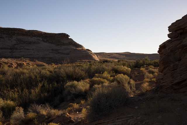 A narrow canyon contains scrub brush and dirt ground, with rock walls rising on either side.