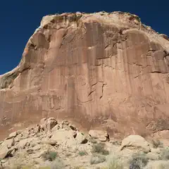 A large rock formation with red and brown hues, featuring a flat top and jagged edges, set against a clear blue sky.