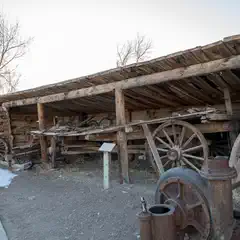 An old wooden shed with a sloping roof is filled with various pieces of rusted machinery and equipment, including large wheels and metal parts.