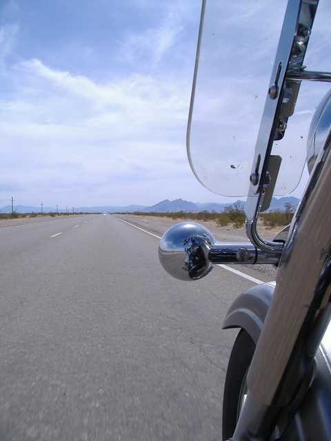 A highway stretches into a distant, mountainous landscape, partially framed by a motorcycle's bodywork.