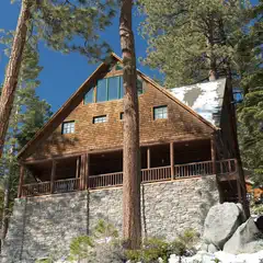 A two-story wooden cabin with a stone base has a covered porch and is surrounded by trees and snow.