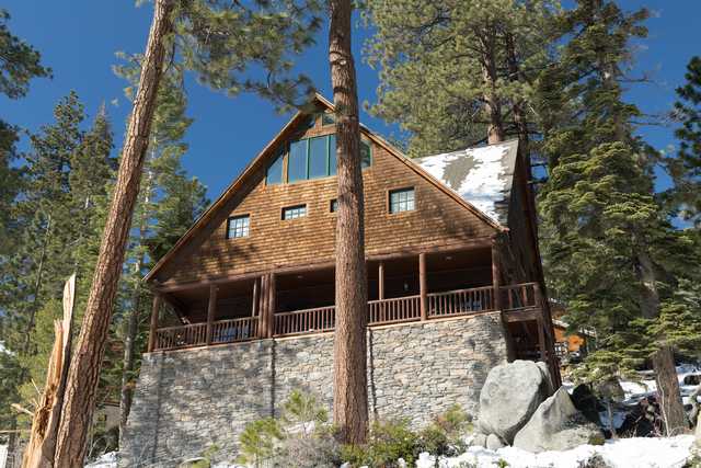 A two-story wooden cabin with a stone base has a covered porch and is surrounded by trees and snow.