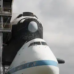 A space shuttle mounted atop a modified Boeing 747, viewed from below with a metal observation structure to the left.