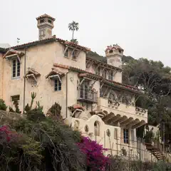 A tan stucco mansion occupies a hillside surrounded by trees and flowering plants.
