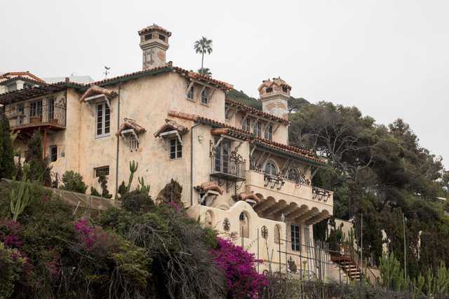 A tan stucco mansion occupies a hillside surrounded by trees and flowering plants.