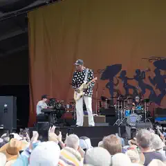 A musician plays an electric guitar on a stage with a band behind him and a crowd in front.