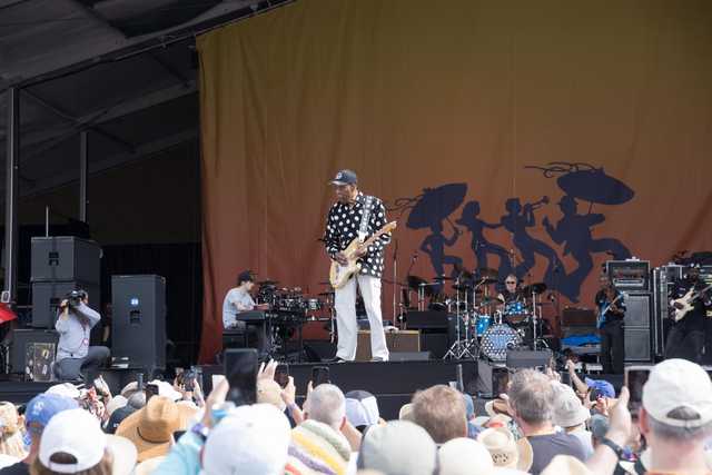 A musician plays an electric guitar on a stage with a band behind him and a crowd in front.