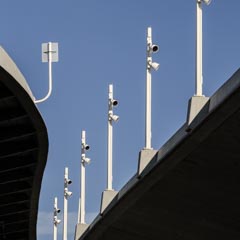 Detail of the underside of a concrete-clad bridge with a row of lamps on white posts rising into a blue sky.