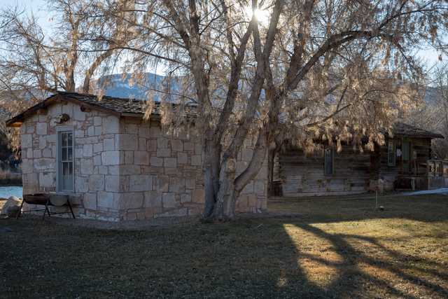 A rustic stone building stands alongside a log cabin, both with brown roofs and surrounded by trees.