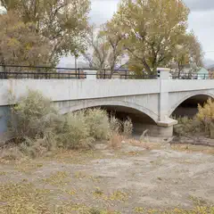 An arched concrete bridge spans a dry riverbed, with trees and shrubs growing on either side.
