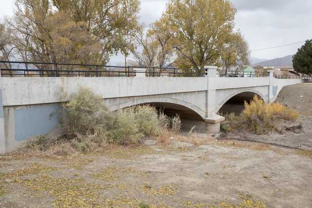 An arched concrete bridge spans a dry riverbed, with trees and shrubs growing on either side.